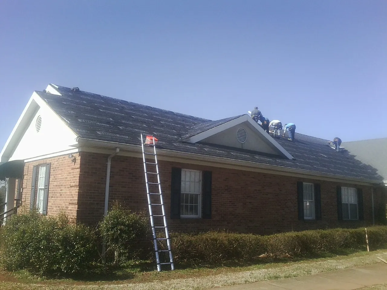 Crew tearing shingles off a brick ranch roof with a ladder set — Metal Roof Repair work by our crew in Grayling