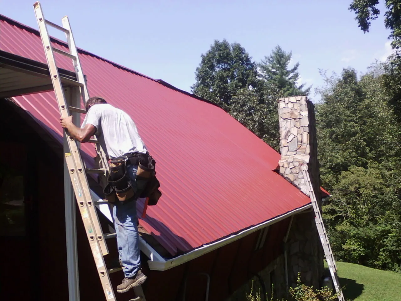 Roofer on a ladder beside a red metal roof and stone chimney — Soffit & Fascia Repair by Roofing Brothers in Grayling